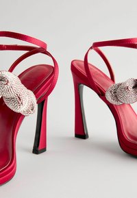 Rote Satin High Heels Sandalen haben einen Strass-Akzent am Riemen, dünne Knöchelriemen und einen kontrastierenden schwarzen Stiletto-Absatz.