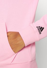 Ręka w przedniej kieszeni różowego sweatshirtu Adidas, pokazująca czarne logo Adidas na prążkowanym mankiecie.