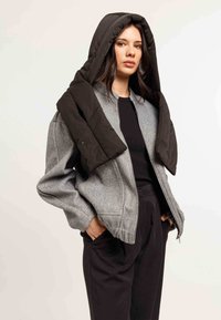 Giacca bomber di lana grigia con cappuccio imbottito nero. Presenta una chiusura con zip, polsini a costine e tasche laterali, indossata sopra una maglia nera.
