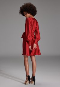 Robe en satin rouge avec une taille ajustée, des manches bouffantes et des accents de boutons. Associée à des chaussures à talons hauts noires, mettant en valeur des jambes lisses et un arrière-plan minimaliste.
