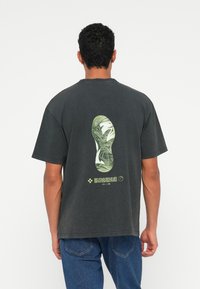 Zwart katoenen T-shirt met een groene grafiek van een schoenafdruk op de achterkant, voorzien van tekst en digitale accenten onder het ontwerp.