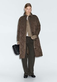 Donna che indossa un lungo cappotto trapuntato marrone sopra un cardigan beige e pantaloni neri, mentre tiene una borsa a pochette nera, in piedi davanti a uno sfondo neutro.