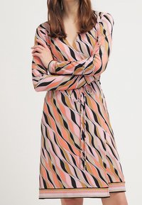 Robe portefeuille à manches longues avec un motif ondulé vibrant en rose, orange, noir et marron. Comprend une ceinture à nouer à la taille et un ourlet droit.