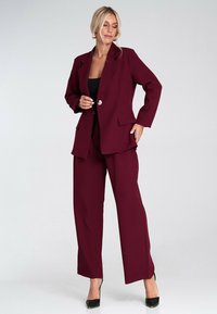 Bordowy dwuczęściowy garnitur z dopasowaną marynarką o wyraźnych klapach i kieszeniach, w połączeniu z szerokimi spodniami. Teksturowana tkanina o subtelnym połysku.
