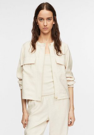 Kobieta z długimi brązowymi włosami nosi jasnobeżową kurtkę na suwak na dopasowanym topie i luźnych spodniach, stojąc na jednolitym białym tle.