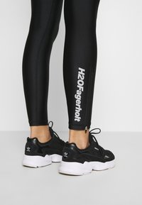 Svarta leggings med dragkedja vid vristen och vit text som säger "H2OFagerholt." Matchas med svartvita löparskor.