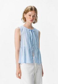 Joven mujer con cabello corto y ondulado lleva una blusa sin mangas de color azul claro con pliegues, de pie frente a un fondo blanco.