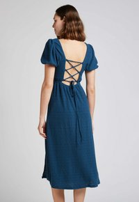 Robe midi texturée bleue avec manches bouffantes, détail de lacet dans le dos et fermeture à nouer. Présente un motif subtil et une coupe décontractée.