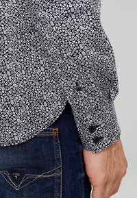 Chemise noire avec un motif floral blanc, munie d'un poignet boutonné et d'une texture lisse, associée à un jean en denim bleu.