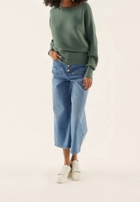 Femme portant un pull côtelé vert, un jean bleu taille haute, jambes larges et raccourci avec des boutons sur le devant, et des baskets blanches, debout sur un fond clair.