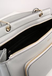 Jasnoszara skórzana torebka z złotymi zamkami, częściowo otwarta, ukazująca czarne wnętrze i zapinaną kieszeń wewnętrzną, z dwiema krótkimi rączkami z złotymi pierścieniami.