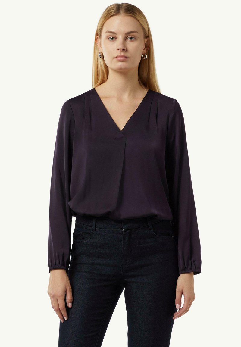 Dunkellila Bluse mit V-Ausschnitt und langen Ärmeln, mit Falten an den Schultern. Kombiniert mit dunklen Jeans aus Denim. Glatte Stofftextur.