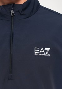 Mornarsko plava jakna na zip s EA7 Emporio Armani logotipom na prsima, djelomično zakopčana do vrata.