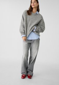 Pullover oversize grigio chiaro in maglia, indossato sopra una camicia a righe blu e bianche, abbinato a jeans svasati grigio sbiadito e scarpe rosse.