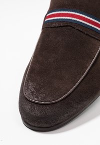 Mocassins en daim marron à texture lisse, avec un bout arrondi et une bande décorative avec des rayures rouges, blanches et bleues.