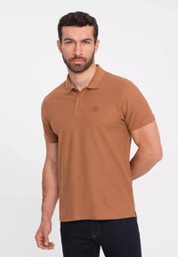 Brun polo-skjorta i bomull, med krage, korta ärmar och en broderad logo på bröstet. Bärs med mörka jeans.