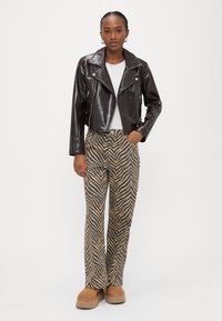 Sieviete, valkājot melnu ādas motociklistu jaku, baltu kreklu, brūnas un melnas zebras raksta bikses un brūnas platformas kurpes, stāvot pret baltu fonu.