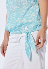 Hellblaue, gemusterte Bluse mit gerafftem Saum und seitlichem Bindeband, getragen über weißen Hosen, aus glattem Stoff und in lässigem Schnitt.