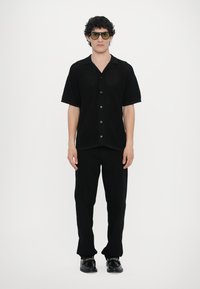 Juodas trumpomis rankovėmis, mygtukais užsegamas marškinis su tekstūrine mezgimo struktūra, derinamas su juodomis kelnėmis ir juodais batais. Modelis dėvi per didelius akinius nuo saulės.