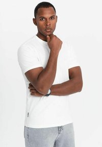 Weißes Baumwoll-T-Shirt mit kurzen Ärmeln, Rundhalsausschnitt und dezenter Logodetails. Kombiniert mit hellgrauen Jeans und einer schwarzen Uhr.