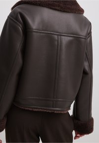 Veste en cuir marron à la coupe courte, dotée d'un col et de poignets en shearling marron doux. Texture lisse avec des détails de couture visibles.