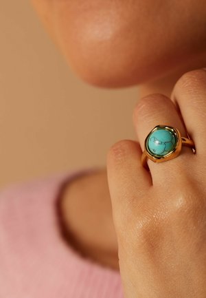 Hand wearing a gold ring with a round turquoise stone, close to face, soft beige background, person in a pink sweater.