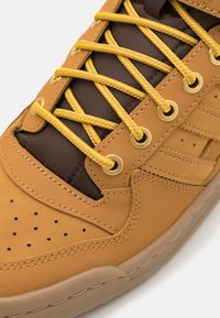 Gros plan sur une sneaker beige avec des lacets jaunes tressés, une languette en cuir brun rembourrée et une semelle texturée beige sur fond blanc.