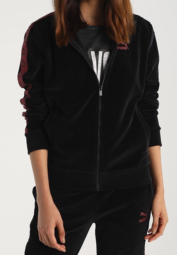 Personne portant un survêtement noir Puma avec des bandes bordeaux sur les manches et le logo sur la poitrine et la cuisse, debout les mains détendues.