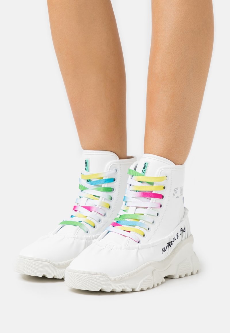 Baskets montantes blanches avec semelles épaisses et lacets arc-en-ciel multicolores portées sur des jambes nues sur un fond uni.