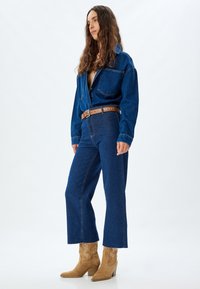Chaqueta de mezclilla y jeans de pierna ancha en azul oscuro, combinados con un cinturón marrón y botas de vaquero en color beige, con un dobladillo deshilachado y material texturizado.