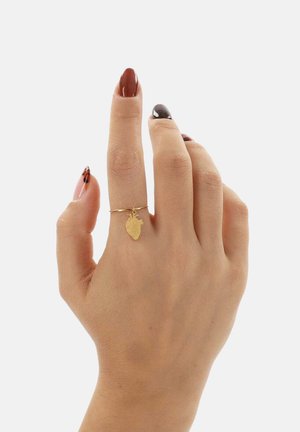 Złoty pierścionek z małym wisiorkiem w kształcie serca, cienka obrączka, noszony na dłoni z zadbanymi paznokciami pomalowanymi na brązowo i czarno.