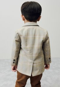 Un jeune enfant portant un blazer à carreaux élégant et un pantalon marron se tient dos au spectateur devant un fond uni.