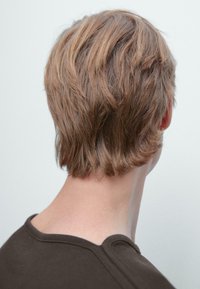 Korte, lichtbruin haar met een golvende textuur en een lichte undercut. De kraag van een donkere top is zichtbaar; de achtergrond is eenvoudig en lichtgekleurd.