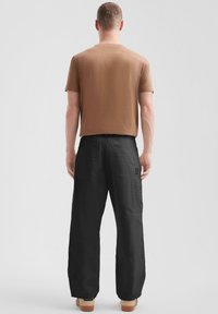 Brązowy t-shirt z krótkim rękawem w połączeniu z luźnymi czarnymi spodniami cargo z dwoma tylnymi kieszeniami, widok z tyłu. Ubranie jest casualowe i swobodne.
