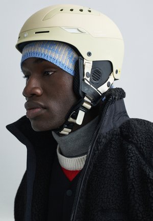 Lysebeige hjelm med justerbare stropper og ventilationshuller, båret over en blå og cremefarvet strikket hue og en struktureret sort jakke.