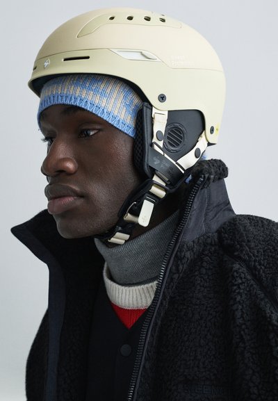 Casco beige chiaro con cinturini regolabili e fori di ventilazione, indossato sopra un berretto a maglia blu e crema e una giacca nera texturizzata.