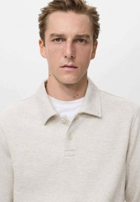 Polo shirt beige chiaro con colletto e tre bottoni. Tessuto liscio e design aderente. Indossato sopra una t-shirt bianca.