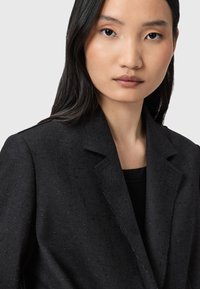 Blazer nero su misura con un tessuto testurizzato, caratterizzato da un colletto a punte e dettagli di cucitura sottili, indossato sopra una camicia nera.