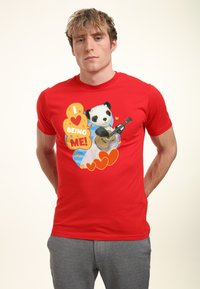 Red short-sleeve t-shirt features a graphic of a panda playing a guitar, with colorful text reading “I LOVE BEING ME!” and heart accents.