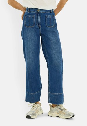 Personne portant un jean bleu à jambes larges avec des poches plaquées devant et des chaussures de sport beiges, debout les mains sur les hanches.