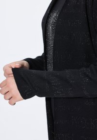 Černý pletený cardigan s volným střihem, se strukturovaným designem a jemnými metalickými akcenty. Rukávy jsou přiléhavé a zakončené manžetou.
