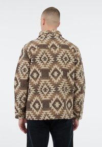 Man in een bruin en beige geometrisch patroonjasje met een kraag, gezien vanuit de rug tegen een witte achtergrond.