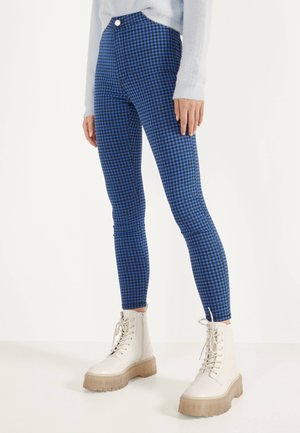 Persona vistiendo pantalones ajustados a cuadros azul y negro y botas blancas con cordones hasta el tobillo, de pie sobre un fondo claro y liso.