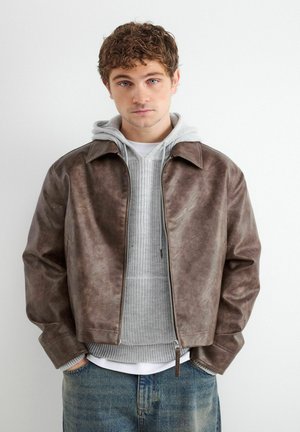 Jeune homme aux cheveux bouclés portant une veste en cuir marron par-dessus un sweat à capuche gris et un jean bleu, debout les mains dans les poches.