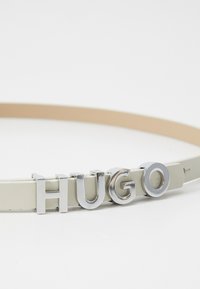 Cintura in pelle beige con grandi lettere argentate che formano la scritta "HUGO", caratterizzata da una texture liscia e un design minimalista.