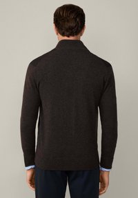 Pull à manches longues marron foncé avec un col haut, tissu texturé et poignets de chemise bleu clair contrastants visibles aux extrémités des manches.