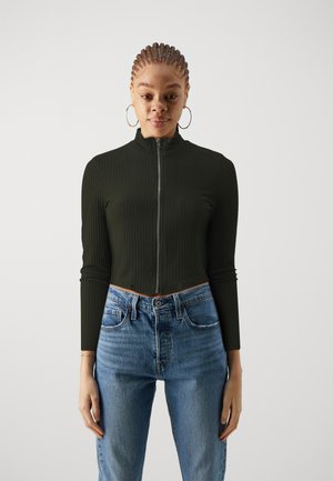 Haut zippé court côtelé vert foncé avec un col haut. Jean en denim bleu clair taille haute avec des coutures visibles et des poches.