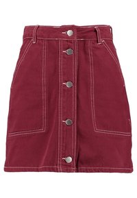 Jupe en denim bordeaux avec un devant à boutons, coutures blanches, deux grandes poches plaquées et une taille ajustée. Silhouette classique en A.
