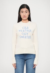 Kreemikas kampsun soonikkoes äärega allosas ja varrukaotstes. Sinise tekstiga, mis ütleb "KÜLMAS ILMAS SOE KAMPSUN." Pehme tekstuur, pikad varrukad.