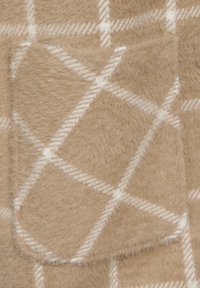 Tela fuzzy beige con un patrón de cuadros blancos, que incluye un pequeño bolsillo frontal y texturas variadas en la superficie.